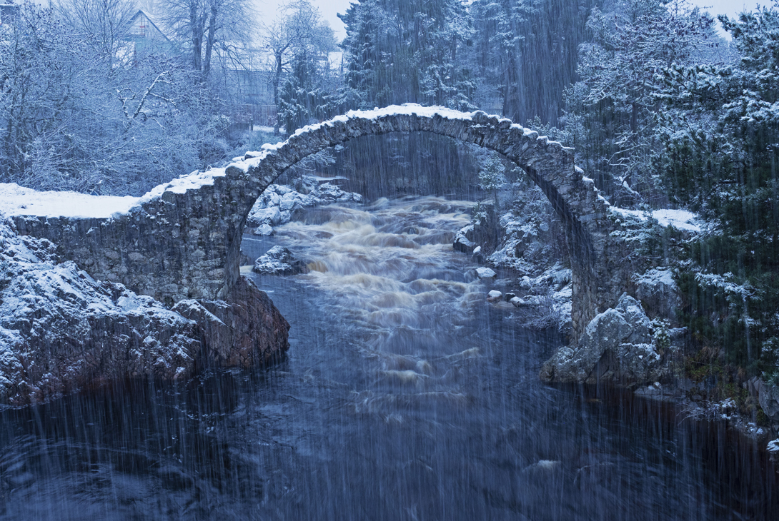 Snow-covered Carrbridge Packhorse Bridge over the rushing Dulnain River, Carrbridge, Cairngorms National Park, Strathspey, Highland Region, Scotland, UK, December